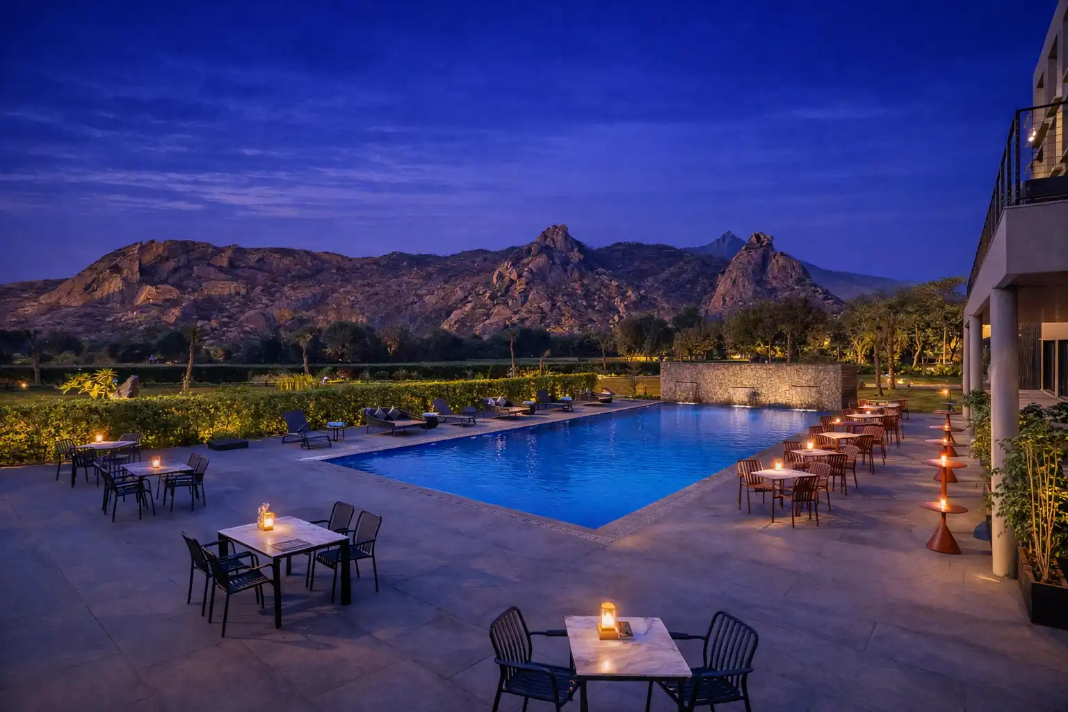 Luxury resort swimming pool and poolside dining tables at Avisa Hotel & Resort near Abu Road under a twilight sky with a backdrop of rugged mountains.